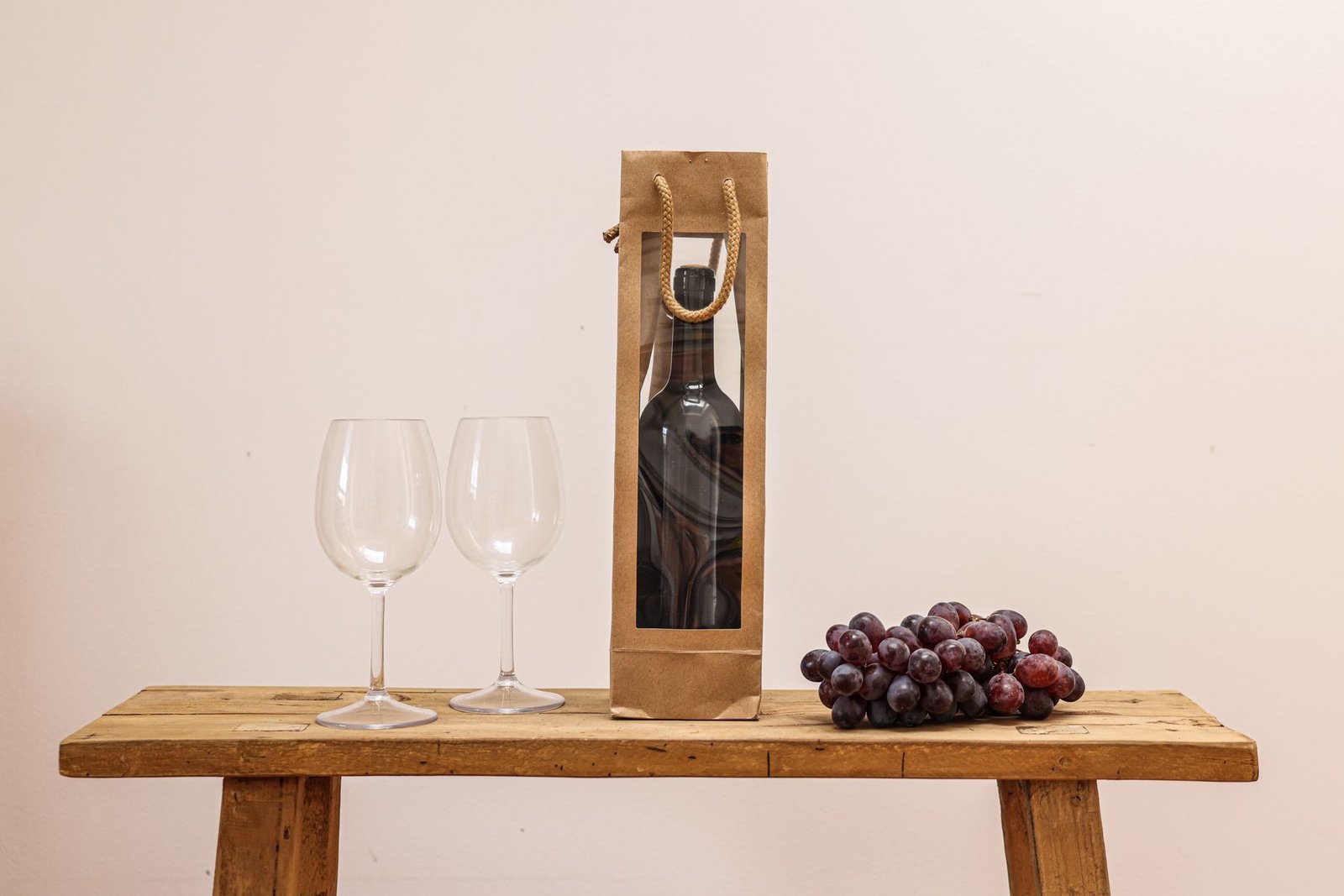A wine bottle in a gift bag beside two glasses and a bunch of grapes on a wooden table, arranged as a wine gift